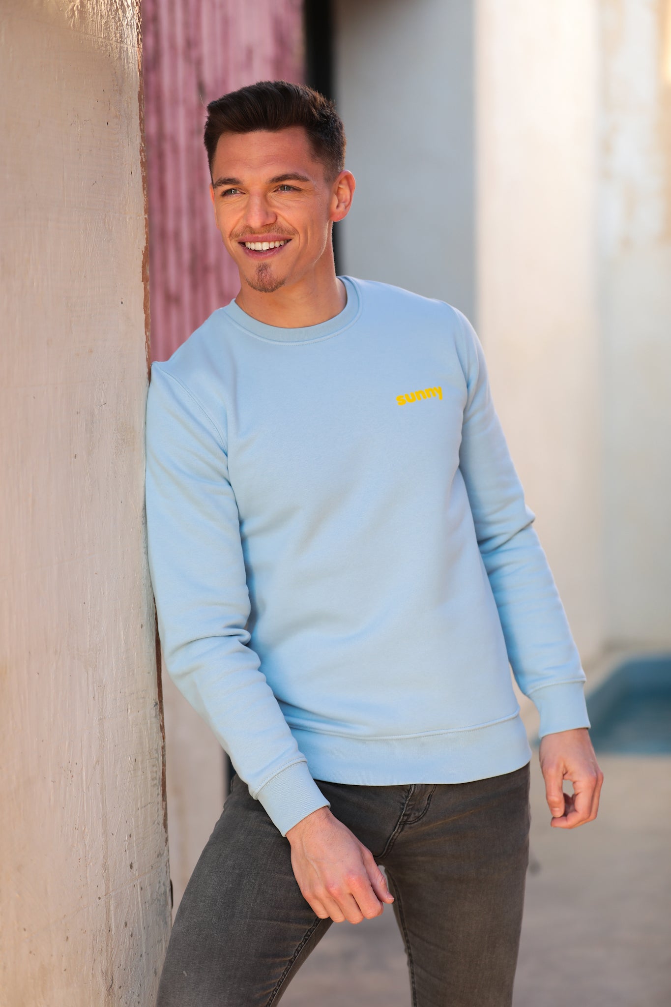 Mannequin homme beau et souriant, pose appuyé contre un mur avec un pull type sweatshirt bleu pastel qui comporte la mention "sunny" en jaune côté coeur. Il a la tête légèrement de profil et regarde avec douceur et émotion vers l'horizon. Un rayon de soleil passe au-dessus de sa tête.
