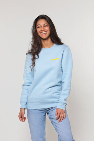 Mannequin femme au sourire éclatant, brune et très belle, pose avec un sweatshirt bleu ciel large et doux sur lequel est inscrit le mot "sunny" en jaune. Elle porte un jean mom bleu pastel.