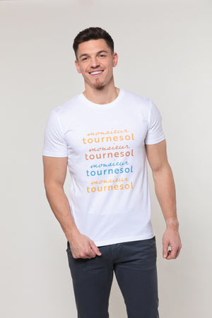 Mannequin homme, brun, de face, souriant, porte un t-shirt blanc avec le logo Monsieur Tournesol décliné en quatre couleurs : orange, brun, bleu et jaune. Il porte un jean bleu gris foncé sur fond gris.