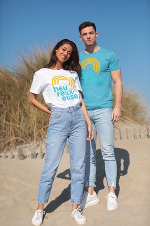 Deux mannequins, un homme et une femme, sur le sable devant une dune, au soleil, portent un t-shirt avec un arc-en-ciel orange sous lequel est écrit heureux·euse en bleu ciel. La femme est à au premier plan, son t-shirt est blanc. L'homme est derrière elle, son t-shirt est de couleur bleu océan.