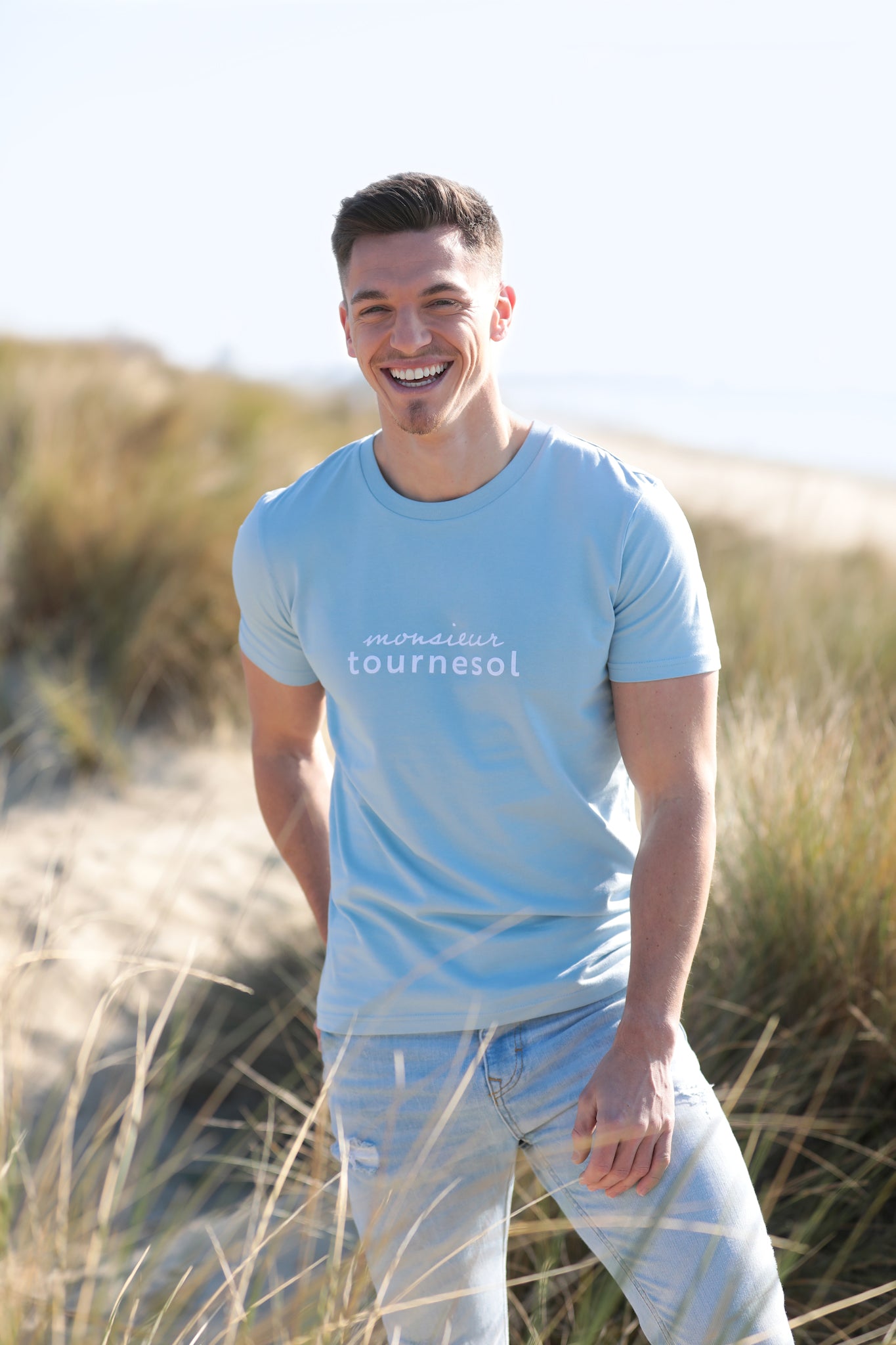 Mannequin homme brun, rit sur une dune de sable devant la mer. Il porte un t-shirt bleu ciel avec inscription Monsieur tournesol en flocage velours blanc et un jean bleu pastel.