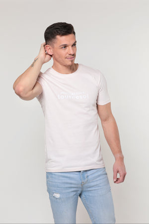 Mannequin homme brun, beau, regard sur le côté et main droite derrière la tête sur fond gris. Il porte un t-shirt shirt rose églantine pastel avec un logo Monsieur Tournesol blanc sur le torse.