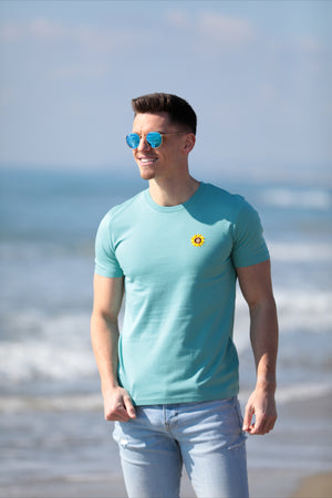 Mannequin homme brun, en bord de mer, porte un t-shirt vert avec une broderie de fleur de tournesol côté coeur et des lunettes de soleil rondes bleues. Il regarde vers l'horizon.